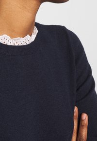 Mörkblå tröja med ribbad halsringning, med en delikat vit spetsdetalj under. Mjuk textur med figursydd passform.