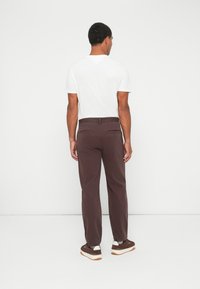 Vīrietis stāv ar muguru, ģērbies baltā T-kreklā, tumši brūnos biksēs un tumši brūnās kedās ar baltiem zole. Fons ir vienkārši balts.