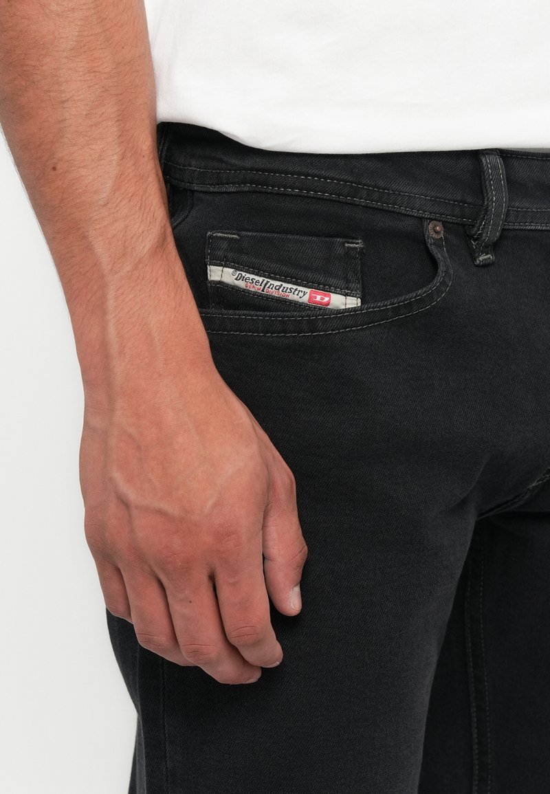 Rechte Hand liegt neben schwarzen Diesel Industry Jeans, die mit einem weißen Oberteil getragen werden, das ein kleines, markiertes Taschen-Detail zeigt.