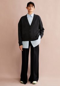 Donkergrijze cardigan met knopen over een lichtblauw overhemd met kraag, gecombineerd met zwarte wijde broek en wit-groene sneakers. Casual look.