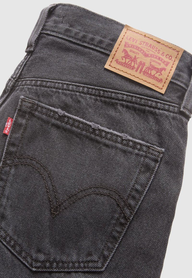 Jeans en denim gris foncé avec une pièce en cuir à l'arrière, présentant des coutures visibles et un design de poche en forme de cœur. Étiquette rouge sur la couture latérale.