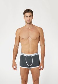 Boxers gris con ribete blanco y el logo "GUESS" en la cinturilla, fabricados con un tejido suave y elástico. El diseño ajustado resalta una silueta estilizada.