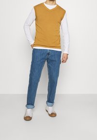 Sennepsgul v-hals ærmeløs sweater over en hvid langærmet skjorte, kombineret med blå jeans og hvide sneakers med brune detaljer.