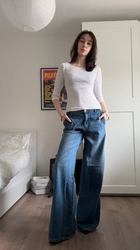 Νεαρή γυναίκα με λευκό μακρυμάνικο τοπ και τζιν παντελόνι με φαρδιά μπατζάκια στέκεται σε υπνοδωμάτιο με τα χέρια στις τσέπες της, πόστερ στον τοίχο πίσω της.