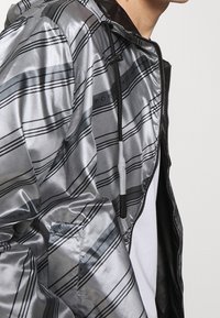 Silver- och svartrandig jacka med huva, som har en glänsande, slät yta och en frontdragkedja. Justerbar dragsko vid huvan.