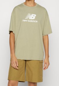 Olivově zelené bavlněné tričko s krátkým rukávem a kulatým výstřihem, s bílým logem "new balance" na hrudi. Spárováno s khaki cargo šortkami.