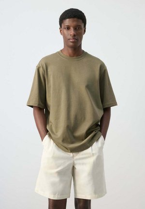 Jeune homme debout, mains dans les poches, portant un t-shirt oversize vert olive et un short écru sur un fond uni.