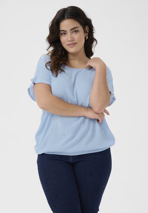 Blouse bleu clair à manches courtes avec une coupe décontractée et un tissu texturé. Portée avec un jean bleu foncé, présentant un col rond et un style décontracté.