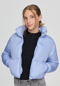 Jasnoniebieska kurtka puchowa z wysokim kołnierzem, pikowanym wzorem, elastycznymi mankietami i zamkiem z przodu. Nosiwana na czarnej bluzce.