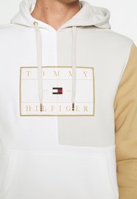 Uomo che indossa un hoodie con blocchi di colore bianco e beige, tasca frontale, cordini e un logo Tommy Hilfiger bordato d'oro sul petto.