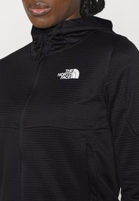 Giacca nera con zip e motivo testurizzato, dotata di colletto alto e un logo bianco de "The North Face" sulla parte sinistra del petto.