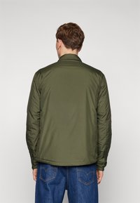 Giacca imbottita verde oliva con colletto classico, dal tessuto liscio e con polsini elasticizzati, indossata sopra pantaloni in denim blu.