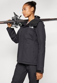 Schwarze wasserdichte Skijacke mit Kapuze, ausgestattet mit einem Reißverschluss vorne und verstellbaren Bündchen; Skifahrer hält Skier auf der Schulter.