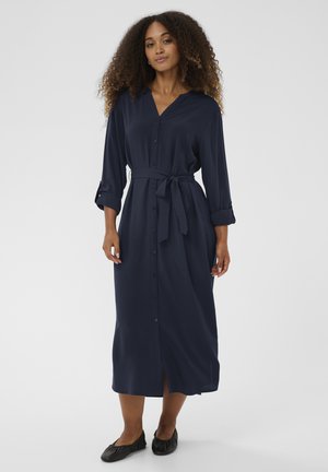 Ženska s kodrastimi lasmi, oblečena v temno modro midi obleko z gumbi, pasom in črnimi ravnimi čevlji, stoji na belem ozadju.