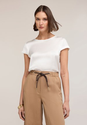 Frau mit kurzen braunen Haaren, die ein weißes Satin-Kurzarmoberteil und hoch taillierte beige Hose mit schwarzem Kordelgürtel trägt.