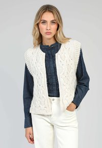 Gilet en ivoire tricoté avec motif en câble et design ouvert à l'avant, associé à une chemise en denim foncé et un pantalon blanc. Look texturé et superposé.