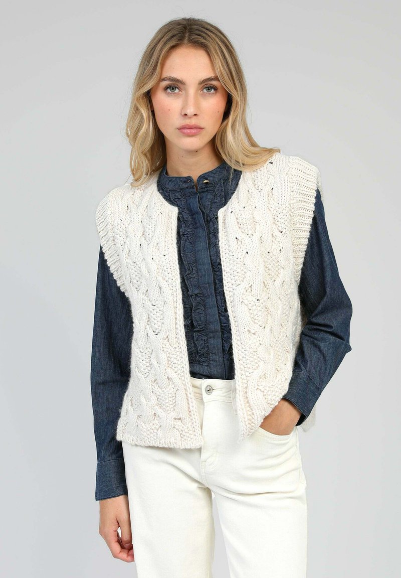 Gilet en ivoire tricoté avec motif en câble et design ouvert à l'avant, associé à une chemise en denim foncé et un pantalon blanc. Look texturé et superposé.