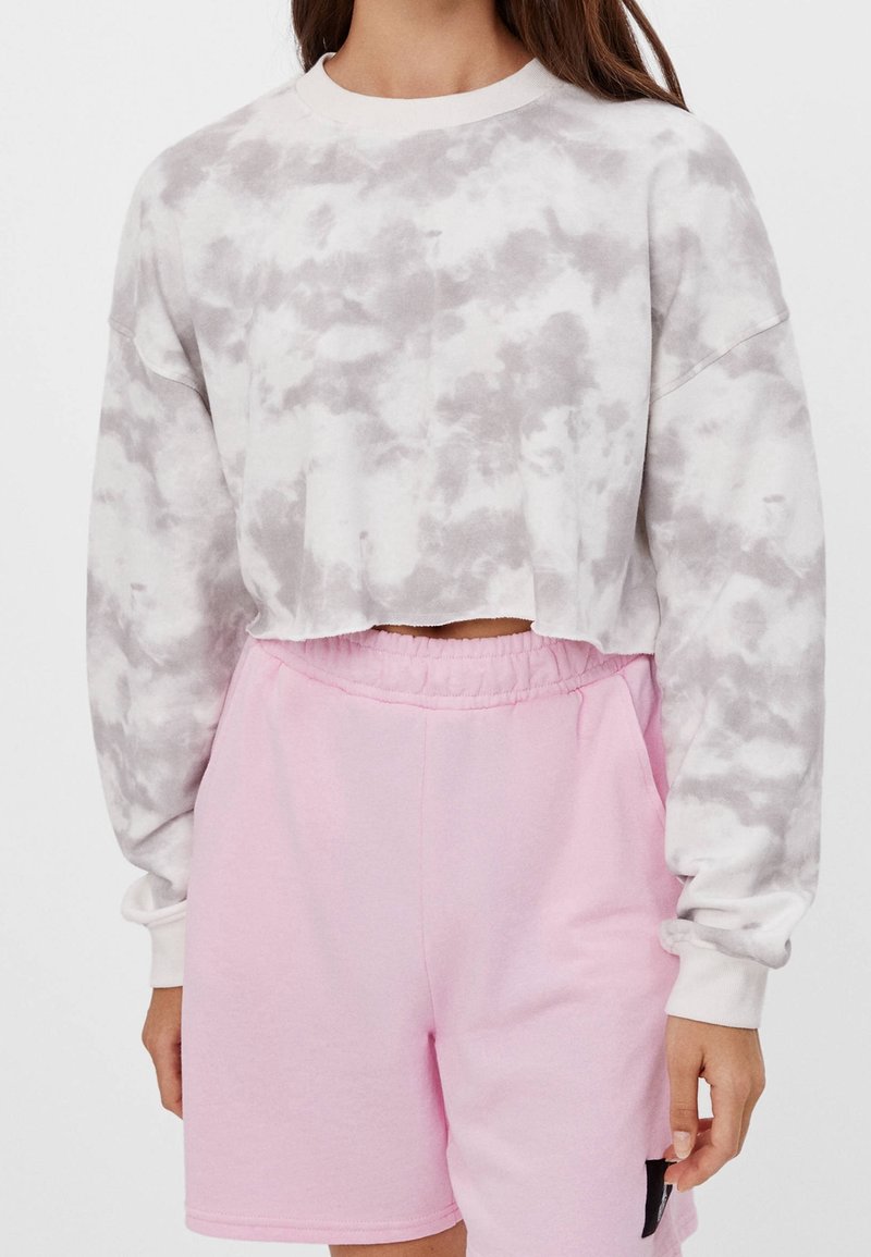 Femme portant un sweat court gris et blanc tie-dye et un short rose clair à taille élastique avec une poche sur le côté droit.