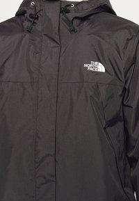 Veste légère noire avec une fermeture éclair à l'avant, une capuche réglable et un logo sur la poitrine gauche. Présente une texture lisse et mate.
