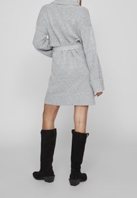 Personne portant une robe en maille gris clair ceinturée et des bottes noires en daim montant jusqu'aux genoux, debout de dos face au spectateur sur un fond uni.