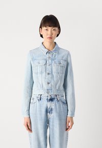 Lys blå denimjakke med en kort design, knappestolpe, doble brystlommer og lange ermer. Har synlige sømdetaljer.