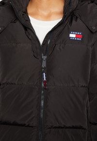 Veste matelassée noire avec fermeture éclair à l'avant, patch logo Tommy Jeans sur la poitrine, portée sur un chemisier blanc côtelé.