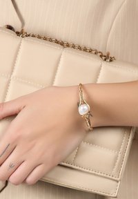Hand with small tattoos wearing a gold bracelet with a large pearl, resting on a beige quilted handbag with a gold chain strap.