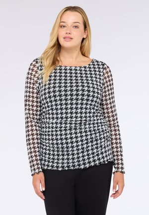 Femme aux cheveux blonds portant un haut à manches longues en pied de poule noir et blanc et un pantalon noir, se tenant devant un fond uni.