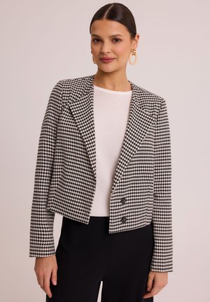 Czarny i biały żakiet w wzór houndstooth o skróconym kroju, z klapami w kształcie litery V i dwoma frontowymi guzikami, noszony na jasnej bluzce.