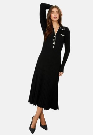 Schwarzes geripptes Kleid mit langen Ärmeln, geknöpftem Ausschnitt, weißen Akzenten, fließender A-Linien-Form und gewelltem Saum. Kombiniert mit schwarzen spitzen Absätzen.