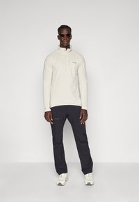 Pullover lungo beige chiaro a maniche lunghe con colletto a zip, in tessuto testurizzato, abbinato a pantaloni neri e sneaker bianche. Indossato occhiali da sole.