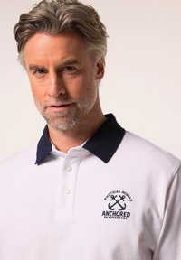 Polo bianco con colletto blu navy, dotato di bottoni e logo a ancora ricamato. Il tessuto sembra liscio con una vestibilità classica.