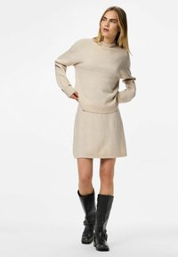 Bežani pleteni pulover s visokim izrezom i rebrastim rukavima, uparen s uskim mini suknjom iste boje. Crne čizme do koljena s kopčama.