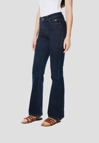 Donkerblauwe uitlopende spijkerbroeken met een hoge taille, voorzien van zilverkleurige hardware accenten en vijf zakken, gecombineerd met bruine sandalen met bandjes.