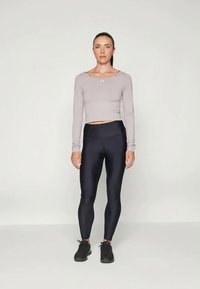Kvinne som står foran og har på seg en lys lilla langermet crop topp, svarte høy midje leggings og svarte treningssko.