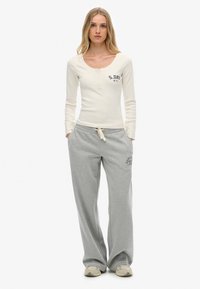 Una mujer de pie con las manos en los bolsillos, vistiendo una blusa larga de manga en color crema y pantalones de chándal gris sueltos, con zapatillas, sobre un fondo blanco.