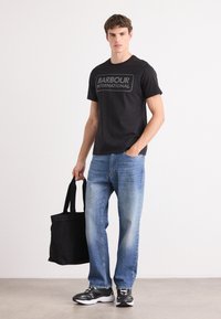 Tricou negru cu logo-ul "BARBOUR INTERNATIONAL", blugi drepti de culoare albastru deschis, geantă mare neagră și pantofi sport negri/albi.