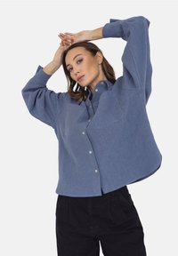 Jasnoniebieska, oversize koszula zapinana na guziki z dwoma przednimi kieszeniami, długimi rękawami i zaokrąglonym dołem. Wykonana z miękkiego, teksturowanego materiału.