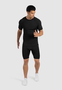 Czarny sportowy top i spodenki, dopasowany krój, krótki rękaw, gładka tekstura, noszone z białymi skarpetkami za kostkę, pozowanie w pozycji stojącej, neutralne tło.