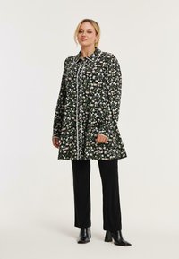 Schwarzes Blumenkleid mit langen Ärmeln, weißen Akzenten und einer Knopfleiste, kombiniert mit schwarzen Hosen und Stiefeln mit Absatz.