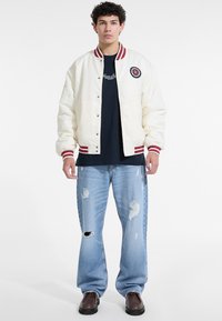 Junger Mann in einer weißen Collegejacke mit roten Streifen, schwarzem Shirt, kaputten hellblauen Jeans und braunen Schuhen, der vor einem weißen Hintergrund steht.