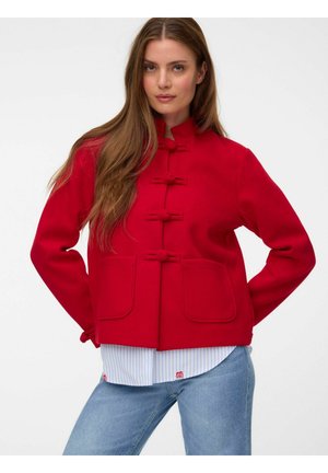 Femme aux longs cheveux bruns portant une veste rouge vif avec des poches avant, des fermetures à bascule, une chemise rayée et un jean bleu sur fond blanc.