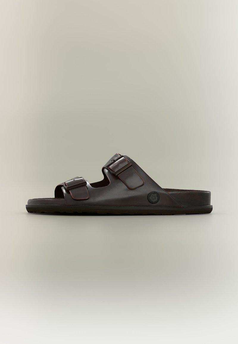 Σανδάλι slide από σκούρο καφέ δέρμα με δύο ρυθμιζόμενα λουριά με αγκράφες και επίπεδη σόλα, εμφανίζεται σε πλευρική όψη σε ουδέτερο φόντο.