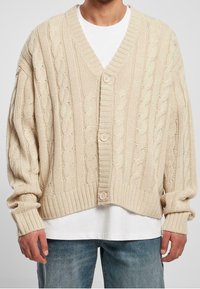 Cárdigan de punto beige con cuello en V, cierre de botones en la parte delantera y puños acanalados. Presenta una textura gruesa y un diseño de corte holgado.