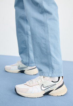 Person iført lyseblå bukser og hvide sneakers med sort og gråt Nike-swoosh, stående på blå gulv.