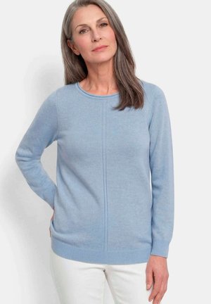 Femme aux longs cheveux gris portant un pull bleu clair à manches longues et un pantalon blanc, debout avec une main sur la hanche.