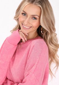 Felpa rosa a maniche lunghe con una texture morbida e polsini a costine. Modella che sorride, indossando orecchini a cerchio colorati e una collana a forma di stella.