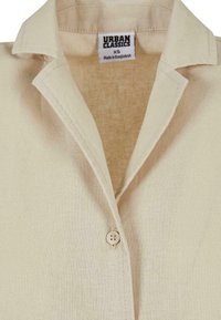 Beige lin skjorte med klippet krave, enkelt knappelukking og en merkelapp som leser "URBAN CLASSICS XS." Strukturerst stoff med synlige sømmer.