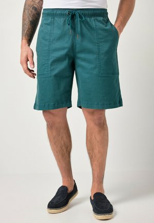 Männliche Person trägt türkisfarbene Shorts mit Kordelzug und großen Fronttaschen, schwarze Schlupfschuhe und ein weißes Hemd, steht vor einem einfarbigen Hintergrund.