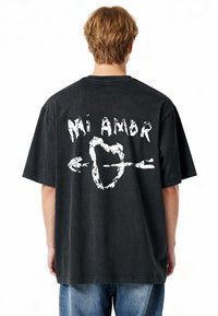 T-shirt noir surdimensionné avec un col rond. Texte graphique blanc "Mi Ambr" au-dessus d'un design de cœur et de flèche au dos. Tissu en coton.